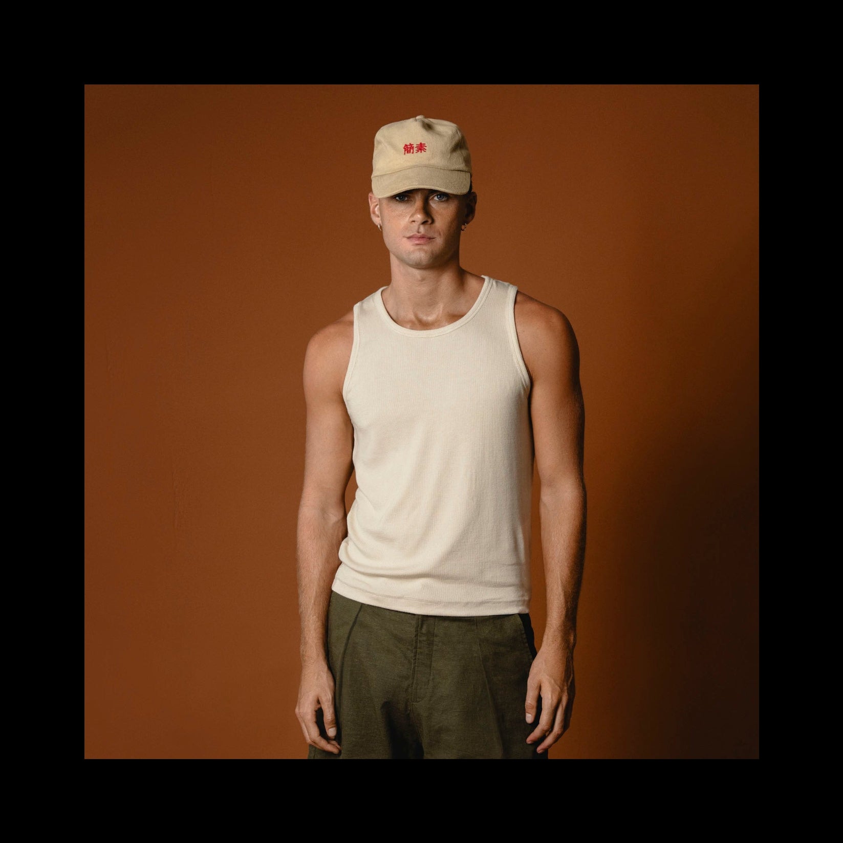 Man wearing a beige tank top and green shorts against a brown background